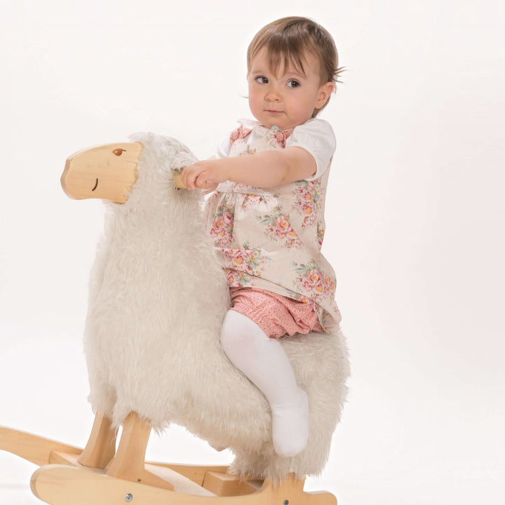 Baby playing on a fluffy rocking sheep, wearing floral top and pink shorts, in a bright and cheerful setting.