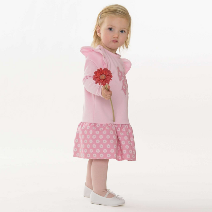 Kleinkind trägt ein rosafarbenes Patternforkids Kleid mit Rüschen und Blumenmuster-Rock, gehalten mit einer Blume.
