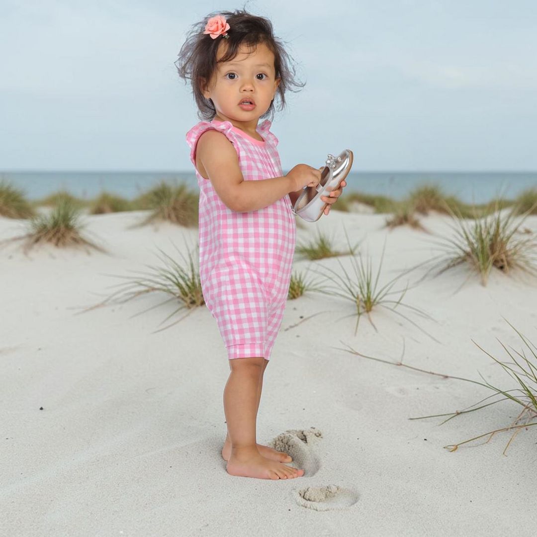 Kleinkind trägt einen rosa-weiß karierten Patternforkids Overall mit Rüschenärmeln am Strand