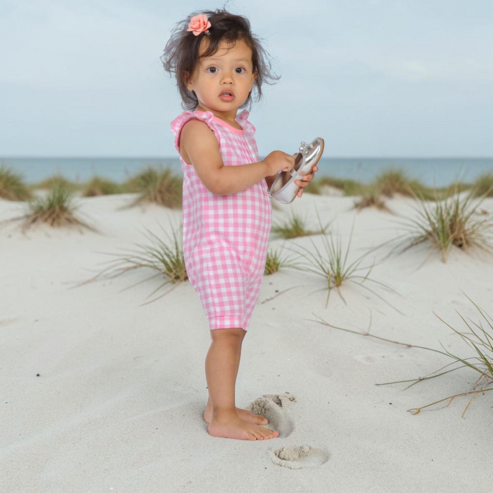 Kleinkind trägt einen rosa-weiß karierten Patternforkids Overall mit Rüschenärmeln am Strand