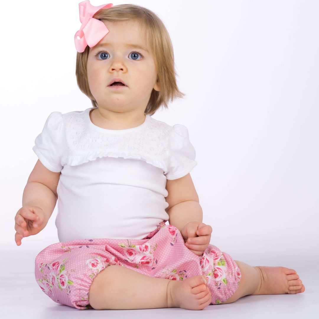 Patternforkids Baby Pumphose Stella aus rosa Stoff mit Blumenmuster, getragen von einem Kleinkind mit rosa Haarschleife