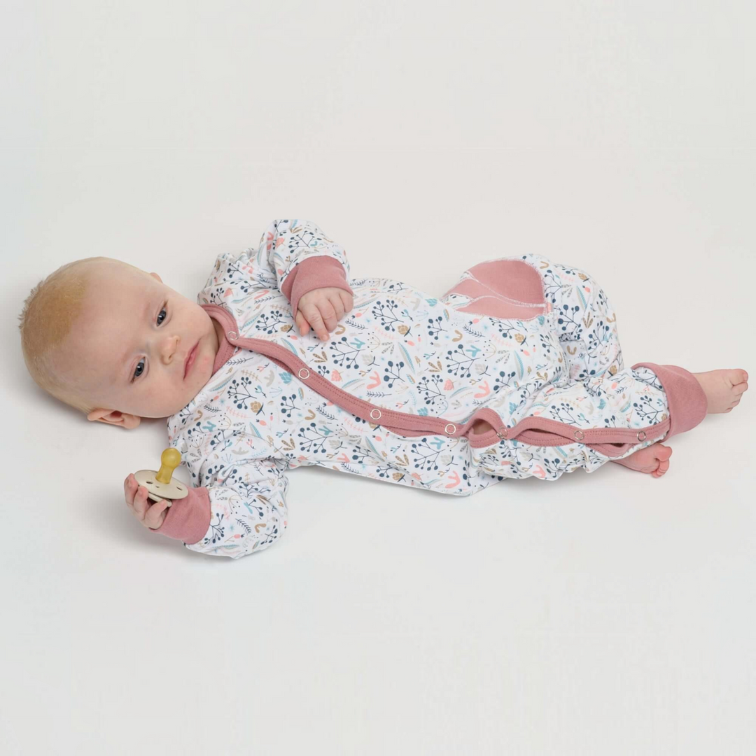 Patternforkids Baby Strampler LISA mit Blumenmuster, Druckknöpfen und rosa Bündchen, getragen von einem Baby.