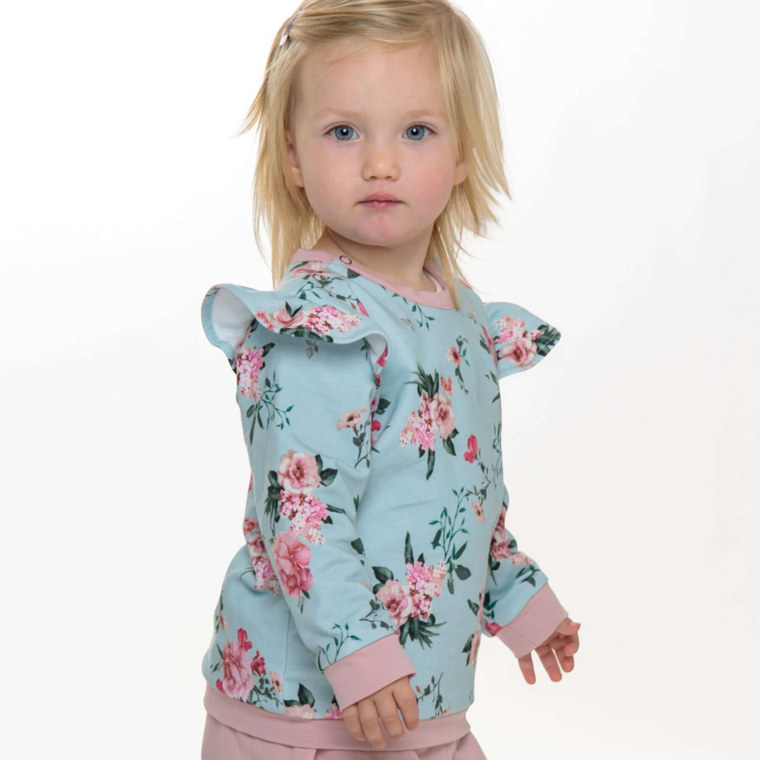 Kleinkind trägt ein hellblaues Patternforkids Sweatshirt mit Blumenmuster, Rüschen an den Schultern und rosa Bündchen.