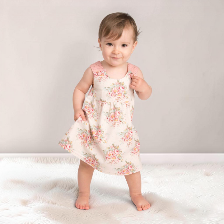 Patternforkids Babykleid Clara mit Blumenmuster und rosa Trägern, getragen von einem kleinen Mädchen auf weißem Fell.