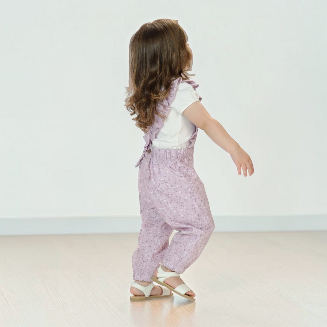 Patternforkids Latzhose DAISY in Flieder mit Rüschen und gekreuzten Trägern, getragen von einem kleinen Mädchen.