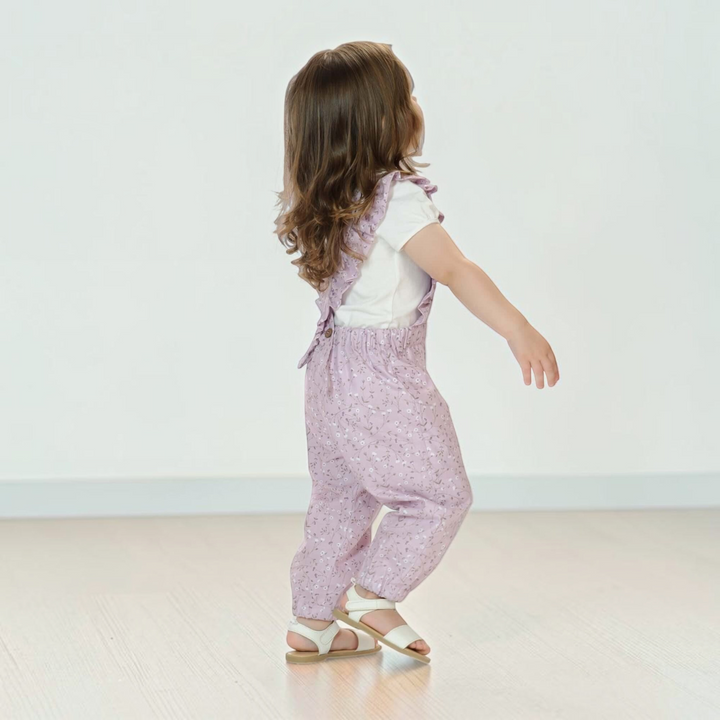 Patternforkids Latzhose DAISY in Flieder mit Rüschen und gekreuzten Trägern, getragen von einem kleinen Mädchen.