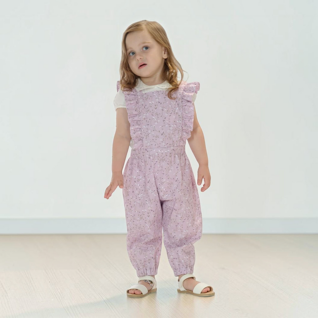 Patternforkids Latzhose DAISY in zartem Rosa mit Blumenmuster, Rüschen-Details und elastischem Bund an einem Mädchen.