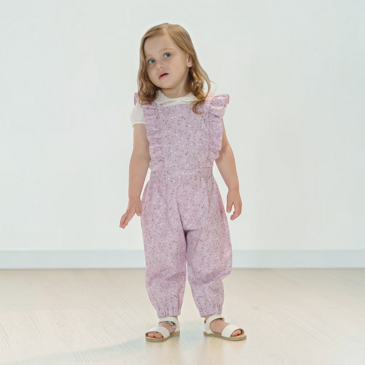 Patternforkids Latzhose DAISY in zartem Rosa mit Blumenmuster, Rüschen-Details und elastischem Bund an einem Mädchen.