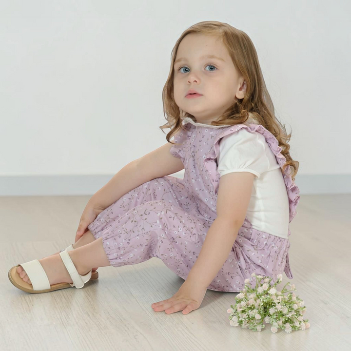 Patternforkids Latzhose Daisy mit Rüschen aus fliederfarbenem Blumenstoff, getragen von einem kleinen Mädchen