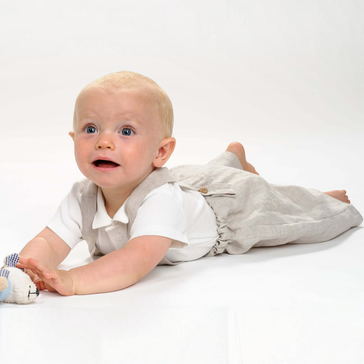 Ein Baby trägt eine beigefarbene Patternforkids Latzhose aus Leinen, liegend auf dem Bauch vor weißem Hintergrund.