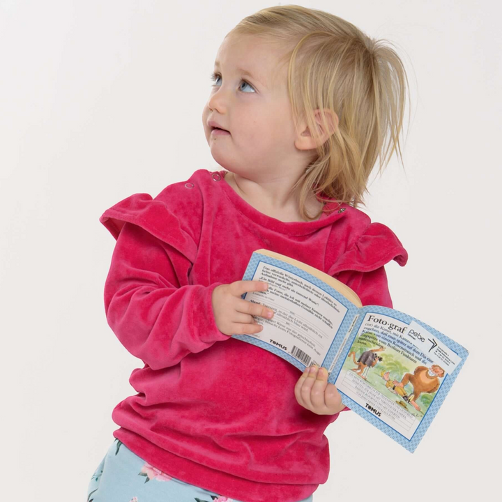 Kleinkind trägt ein pinkes Patternforkids Sweatshirt mit Rüschen an den Schultern und Schulterknöpfen beim Lesen.