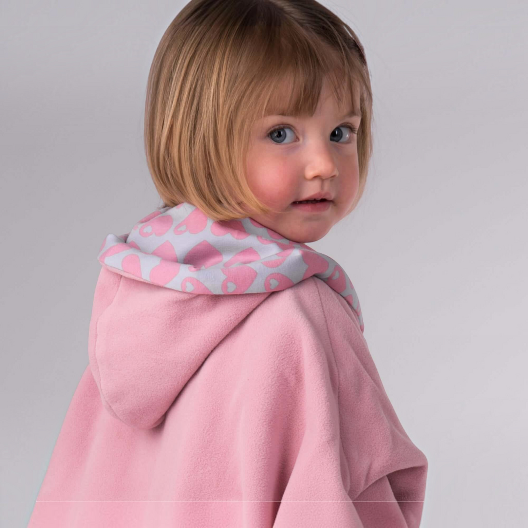 Patternforkids Schnittmuster Set MARA FIOCCO: Kind trägt einen rosa Fleece-Poncho mit Kapuze und Herz-Innenfutter.