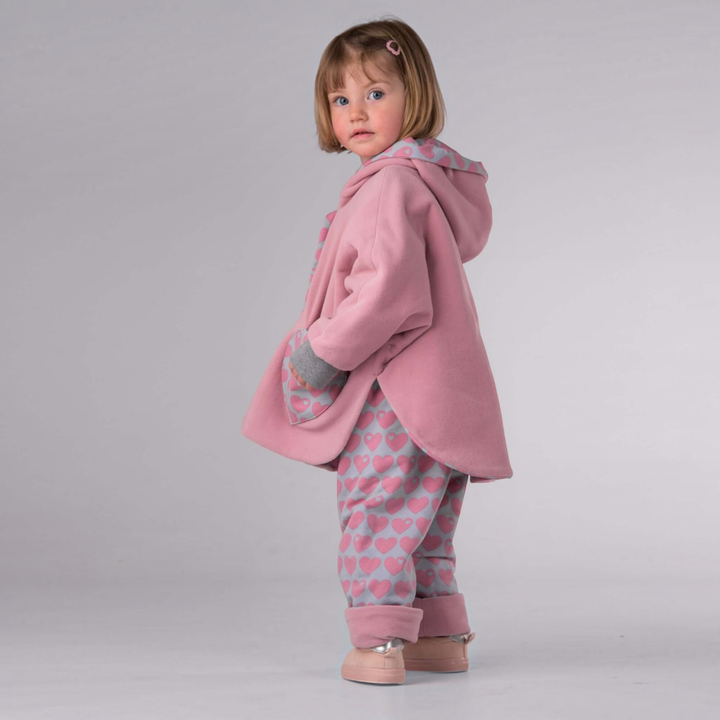 Kind trägt Patternforkids Poncho MARA in Rosa und Wendehose FIOCCO mit Herzmuster, von hinten fotografiert.