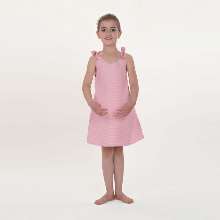 Mädchen trägt ein rosa kariertes A-Linien Kleid von Patternforkids mit Schleifen an den Trägern vor weißem Hintergrund