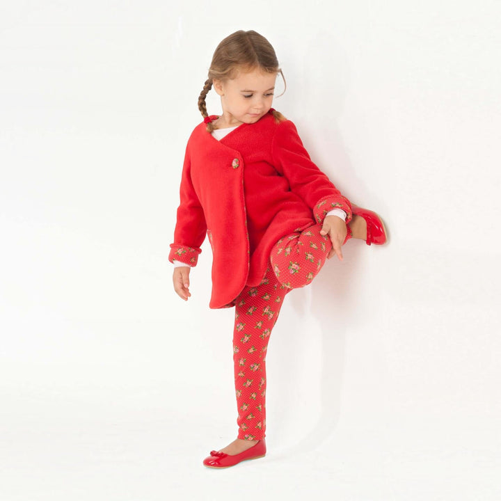 Mädchen trägt rotes Wickelkleid und leggings mit Blumenmuster, posiert spielerisch, zeigt Bein, fröhliche Kinderkleidung.